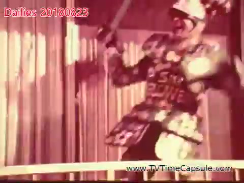 The still shows a performer dressed in an elaborate costume, wielding a large object that resembles a sword. The setting appears to be a stage or entertainment venue, with a backdrop that suggests a theatrical performance. The visual style is reminiscent of vintage film, indicating that it was likely captured on 16mm film stock. The performer exudes an energetic or dramatic presence, contributing to the sense of action.