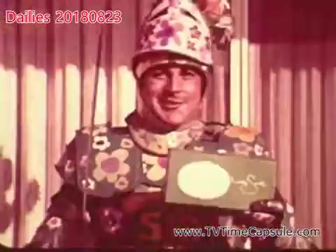 The still depicts a man dressed in a colorful, flower-patterned costume and a whimsical hat, smiling while holding a green card with a white oval on it. The vibrant colors and playful attire suggest a lighthearted or comedic context, possibly from a children's show or a theatrical performance. The overall mood appears cheerful and festive.