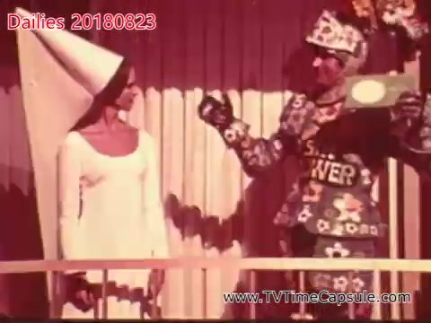 The still features two characters: a woman in a simple white dress and cone hat, and a man dressed in a colorful, patterned costume. The scene appears to be playful or whimsical, possibly from a performance or television segment. The background has a striped design, suggesting a lighthearted atmosphere. The man is gesturing towards the woman, who looks engaged in the moment.