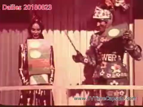 The still features two performers, a woman in a flowing outfit holding a sign and a man in a colorful, festive costume holding a pointer. They appear to be in a celebratory setting, possibly a game show or promotional event, with an engaging atmosphere. The backdrop suggests an outdoor or festive theme.