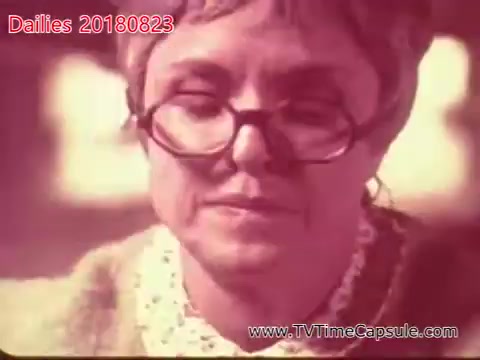 The still features an older woman with glasses and a thoughtful expression. She appears to be wearing a light-colored blouse or dress, with details implying a homey or vintage style. The lighting gives the image a warm, nostalgic quality.