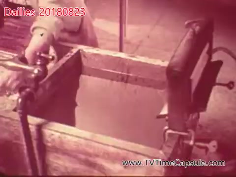 The still features a wooden water trough, possibly used in a workshop or industrial setting. A person's hand is visible, interacting with a faucet or valve over the trough. The image has a reddish tint, suggesting an older film aesthetic, likely from a 16mm reel. The context hints at daily activities or tasks related to a specific project or environment.