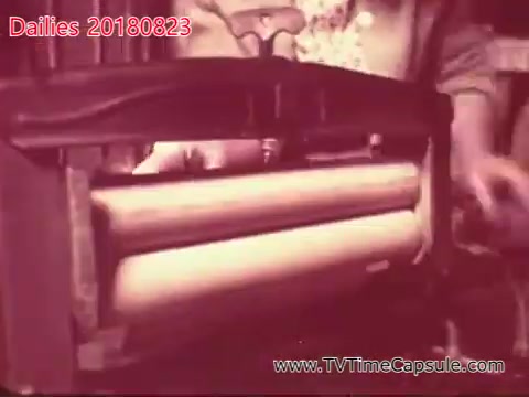 The still depicts a vintage printing press or similar machinery, focusing on rolls of paper being processed. The image captures the mechanical components and the operator's hands, suggesting a historical context related to printing or production. The sepia tone gives it a nostalgic feel, indicative of earlier film technology.