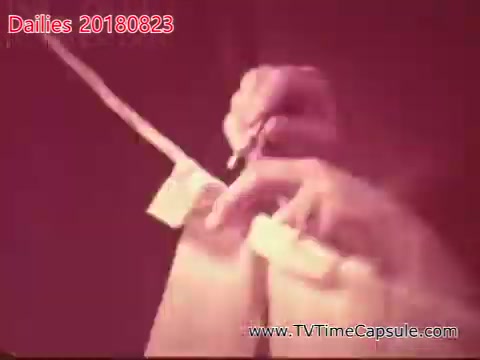 The still depicts a close-up of hands performing an action related to a film or project, possibly handling equipment or making notes. The image has a vintage quality, likely due to its 16mm format, and captures a moment of creativity or production, suggesting an intimate behind-the-scenes look at the filmmaking process. The colors are muted, adding to the nostalgic tone.