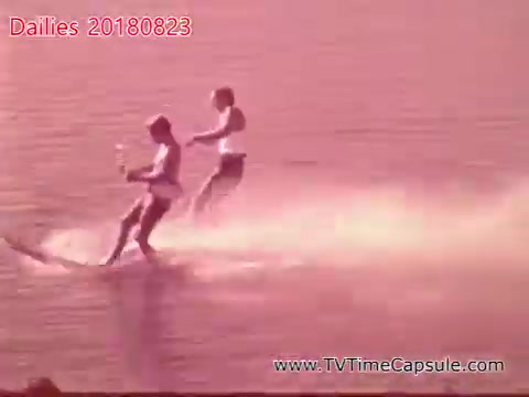 The still depicts two individuals water skiing on a calm body of water, showcasing a cheerful outdoor activity. The scene is bathed in a warm, pinkish hue, indicative of the lighting conditions during filming. The skiers appear to be enjoying themselves, suggesting a fun, recreational atmosphere.