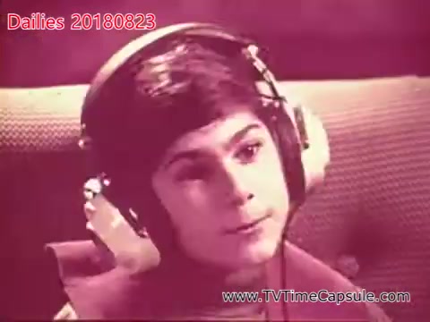 The still shows a young boy wearing large headphones, looking thoughtfully off to the side. The background is blurred with a soft focus, suggesting a recording or listening scenario. The overall tone appears vintage, likely due to the 16mm film format.