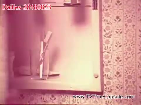 The still features a bathroom setting with a cabinet partially open. Inside, there are various items, including a toothbrush placed in a glass and possibly towels. The background is adorned with patterned wallpaper, contributing to a nostalgic atmosphere typical of vintage home scenes. The overall tone suggests a focus on everyday life and domestic details.
