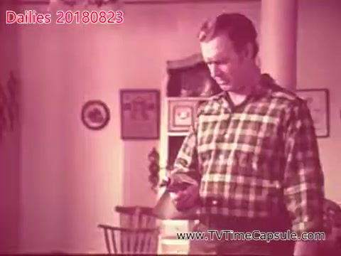 The still from 'dailies 20180823-scott-towels-etc' features a man in a plaid shirt, looking intently at an object in his hands. The setting appears to be a cozy, modest interior, with wooden furniture and decorative items on the walls. The color tone gives a nostalgic feel, suggesting it may be from an earlier era. The man's expression indicates concentration or concern, hinting at a significant moment in the narrative.