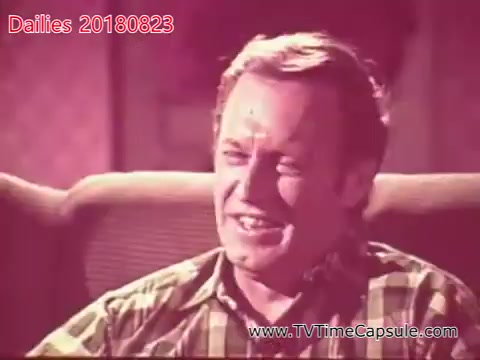 The still captures a middle-aged man with a cheerful expression, sitting comfortably on a couch. He appears to be engaged in conversation, likely sharing a lighthearted moment. The overall tone of the image suggests a casual and friendly atmosphere.