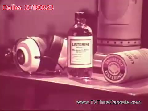 The still features a vintage setup with a bottle of Listerine prominently displayed in the center. To its left, there's a pair of headphones, and to its right, a cylindrical container, also associated with Listerine. The composition suggests a thematic focus on personal care or hygiene, typical of advertising or promotional material. The overall aesthetic reflects an older film style, likely produced for dailies or test screenings.