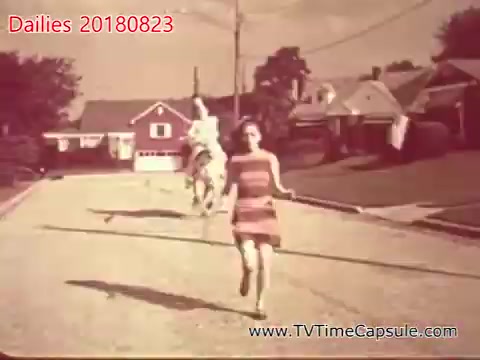 The still captures a nostalgic scene on a suburban street. A young girl wearing a striped dress runs joyfully, while a horse, ridden by another individual in the background, trots along. The setting includes modest homes and a tree-lined backdrop, enhancing the vintage feel of the moment.