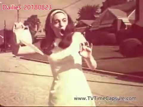 The still from 'dailies 20180823-scott-towels-etc' features a woman in a white dress and headband, striking a playful pose in a suburban setting. She appears to be expressing surprise or excitement, holding something in her hand, with a street and houses visible in the background. The vintage aesthetic suggests it might be from an earlier era of film.