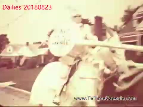 The still features a figure dressed in light-colored attire, riding a horse and holding a shield. The scene appears to be outdoors, likely depicting a festive or historical event. The image has a vintage, sepia tone, suggesting it may be from an earlier era or styled to evoke nostalgia.