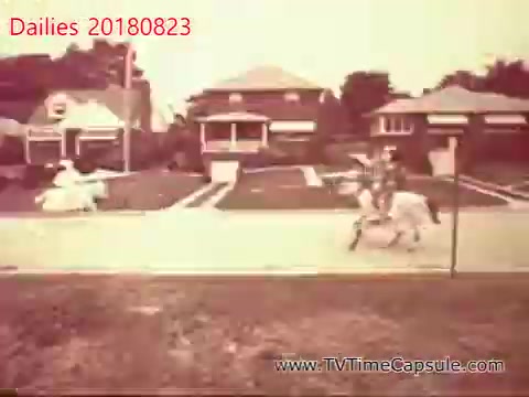 The still captures a vintage scene featuring a person on horseback riding along a suburban street, with houses lining the background. The overall tone is sepia, suggesting a nostalgic or historical context. The scene evokes a sense of simplicity and leisure in a past era.