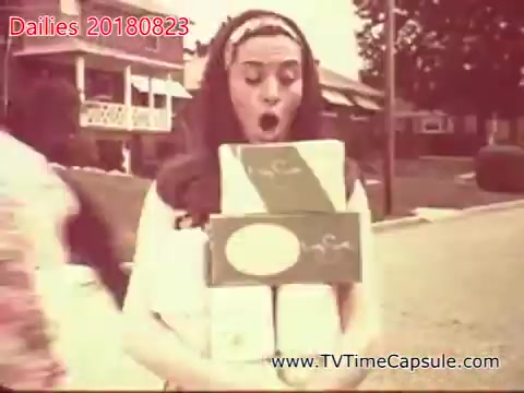 The still depicts a woman outdoors, appearing surprised or excited as she balances several boxes in her arms. The setting suggests a suburban neighborhood, with houses visible in the background. The image has a vintage aesthetic, typical of 16mm film.