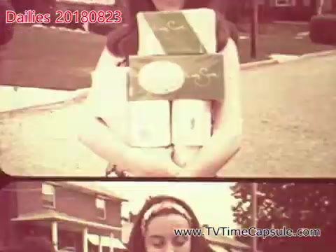 The still features a young girl standing outdoors, holding several packages wrapped in green and white. She appears to be posing for the camera on a residential street, with houses and trees visible in the background. The image has a vintage look, typical of 16mm film, presenting a nostalgic atmosphere.