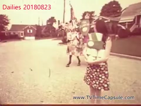 The still from 'dailies 20180823-scott-towels-etc' features a street scene from a bygone era. A girl in a polka dot dress is holding a small package, while a person in a colorful costume, possibly a performer or mascot, walks in the background. The setting appears to be suburban, with houses lining the street, creating a nostalgic atmosphere.