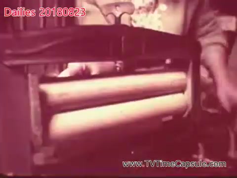 The still features a close-up of a mechanical device, likely a printing press or similar machinery, showcasing its cylindrical rollers. A hand is seen adjusting or interacting with the equipment, indicating an operational context. The overall atmosphere appears vintage, consistent with an older film style.