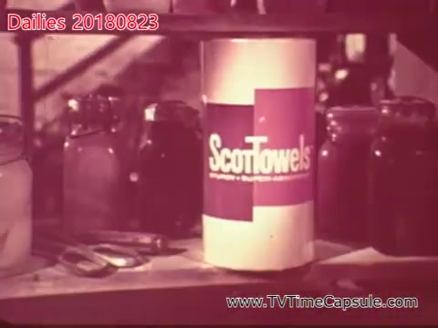 The still features a cylindrical container of ScotTowels prominently displayed on a shelf, surrounded by various jars and utensils. The image likely captures a moment focused on household or kitchen products, suggesting a promotional or advertising context. The color palette and style indicate a retro aesthetic typical of the late 20th century.