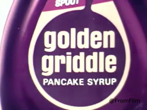 The still features a close-up of a bottle of Golden Griddle Pancake Syrup from the 1970s. The label prominently displays the brand name 'golden griddle' in bold, playful lettering, set against a purple background. There's a focus on the bottle's spout, giving it a classic, retro feel typical of advertisements from that era. The overall presentation evokes a sense of nostalgia associated with breakfast products from the 70s.