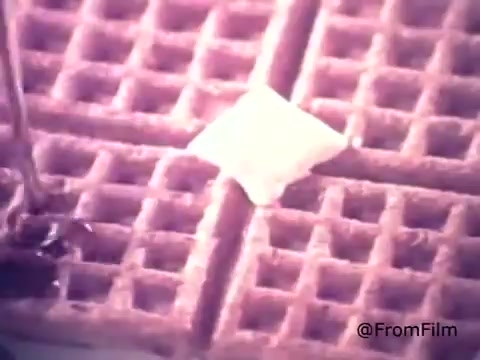 The still from the 1970s Golden Griddle Pancake Syrup commercial features a close-up of a waffle topped with a square of melting butter. A stream of syrup is being poured over the waffle, emphasizing its rich, sweet texture. The scene captures the nostalgic and indulgent nature of breakfast with Golden Griddle syrup. The overall tone is warm and inviting, typical of food advertisements from that era.