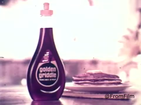 The still from the 1970s Golden Griddle Pancake Syrup advertisement features a bottle of syrup prominently displayed in the foreground. The bottle, designed with a distinctive silhouette and an eye-catching label, reflects the branding of the era. In the background, a stack of pancakes sits on a plate, showcasing a delicious meal ready to be enjoyed. The color palette is warm, evoking a nostalgic feel typical of vintage advertisements.