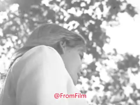 The still from 'Camp Fire Girls PSA - 1969' features a young girl looking upwards, likely outdoors in a natural setting. The image is black and white with a soft focus, capturing sunlight filtering through tree leaves. The girl's expression seems contemplative, aligning with the PSA's themes of community and personal growth. The visual quality appears slightly warped, potentially affecting clarity.