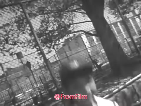 The still from 'Camp Fire Girls PSA - 1969' presents a black-and-white frame with a slightly warped quality, likely affecting both visual clarity and soundtrack. The scene seems to depict a young person in a casual outdoor setting, possibly near a fence or park. The environment includes trees and buildings in the background, suggesting a community space. Overall, the image reflects the style and themes typical of public service announcements from that era.