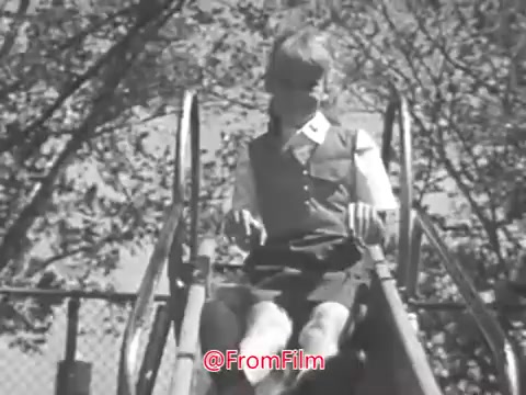 The still from the 'Camp Fire Girls PSA - 1969' features a young child seated on a slide, preparing to go down. The image is slightly warped, reflecting the vintage quality of 16mm film. The background consists of trees, indicating an outdoor setting, likely a playground. The child is dressed in a shirt and shorts, indicating a casual, playful atmosphere.
