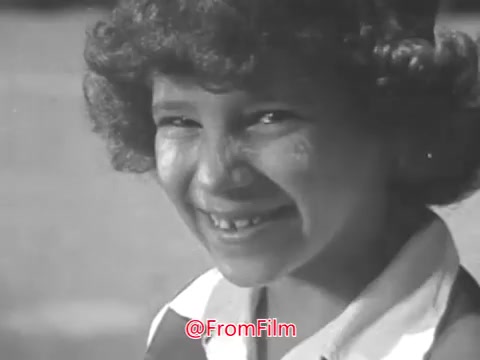 The still from the 1969 'Camp Fire Girls PSA' features a young girl with curly hair smiling at the camera. The image has a vintage aesthetic, characteristic of 16mm film, and appears slightly warped. The girl's expression conveys a sense of joy and engagement, fitting the positive message typically associated with public service announcements from that era. The soundtrack is suggested to have some distortion, aligning with the film's age.