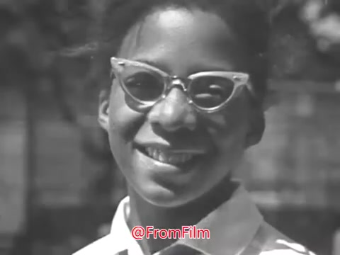 The still from 'Camp Fire Girls PSA - 1969' features a young girl with glasses smiling at the camera. The image likely exhibits the characteristics of 16mm film, which may include a slight warp or graininess. The background suggests an outdoor setting, enhancing the cheerful atmosphere of the moment captured.