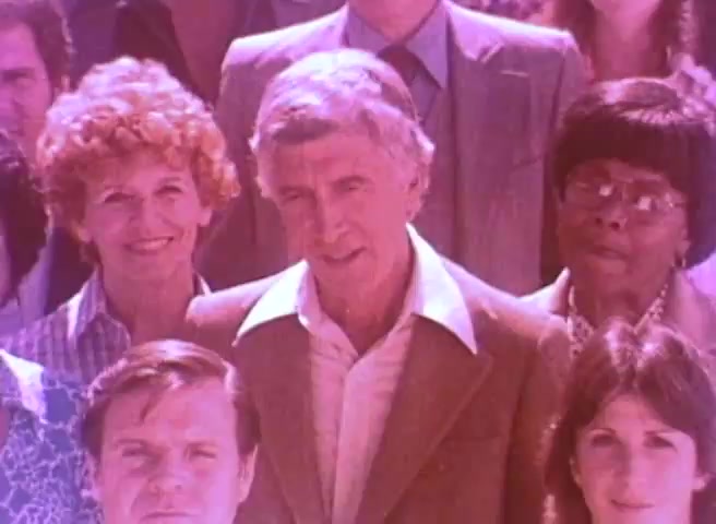 The still features a diverse group of people gathered together, with a focus on a prominent individual in the center. The scene has a vintage aesthetic characteristic of a 16mm print, possibly showing Kirk Douglas directing a message related to the 1980 Census. The background includes individuals with varying expressions, creating a sense of community and engagement in the public service announcement.