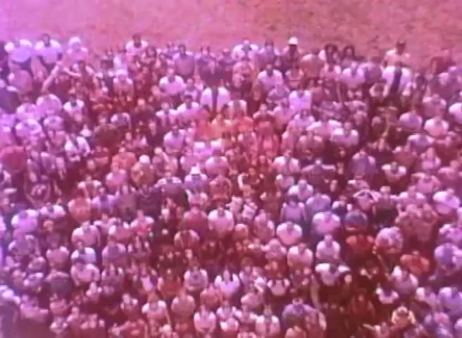 The still features a large, diverse crowd gathered together, viewed from above. The scene captures a lively atmosphere, conveying a sense of community and unity. The colors appear slightly faded, typical of a 16mm print, adding a nostalgic quality to the image. The context is related to a public service announcement promoting the 1980 Census, emphasizing the importance of participation.