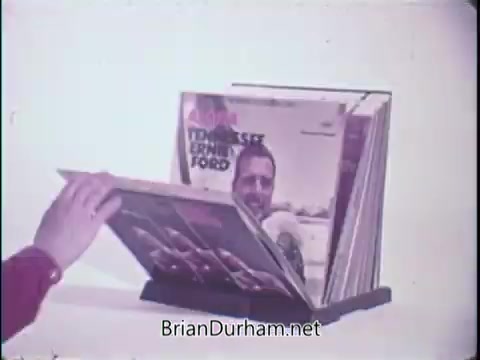 The still features a hand flipping through a record album holder displaying vinyl records. One of the visible covers prominently features Tennessee Ernie Ford. The setting emphasizes a retro, vintage aesthetic typical of late 20th-century advertisements, especially focused on home entertainment products. The scene suggests convenience and accessibility in music selection, aligning with the tagline promoting the Record Matic Automatic Record Selector.