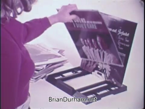 The still features a woman interacting with the 'Record Matic Automatic Record Selector,' a vintage promotional product. She appears to be handling a record, which is part of the selector system designed to automate music playback. The scene captures a nostalgic 1970s home environment, highlighting the convenience of music selection with the device. The packaging reflects a retro design aesthetic typical of products marketed in that era.