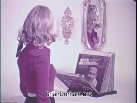 The still features a woman with wavy blonde hair, dressed in a red top, looking at herself in a mirror while holding a record album. In the foreground, a stack of vinyl records can be seen. The mirrored reflection adds a layer of nostalgia, fitting the theme of vintage music. The image captures a moment indicative of the 1970s, showcasing the excitement around home entertainment.