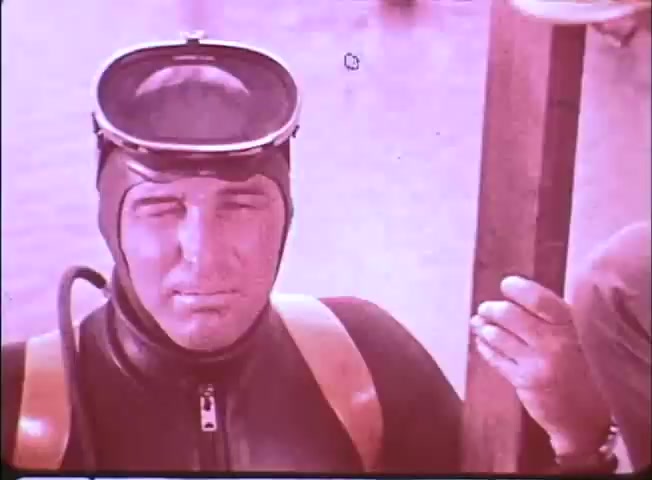 The still features a diver in a vintage diving suit, complete with a mask and snorkel. He appears focused and prepared for an underwater adventure. The background suggests a marine environment, characteristic of exploration themes present in the trailer for 'Around the World Under the Sea.' The color palette has a nostalgic tone, reflecting the era of the film.