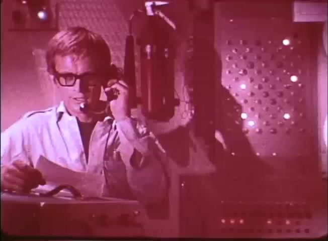 The still features a young male character in a futuristic setting, wearing glasses and a headset, engaged in communication. He's surrounded by technological equipment, suggesting a high-tech environment, likely aboard a submarine. The scene has a reddish tint, creating a dramatic and adventurous atmosphere indicative of the underwater exploration theme of the trailer for 'Around the World Under the Sea.'