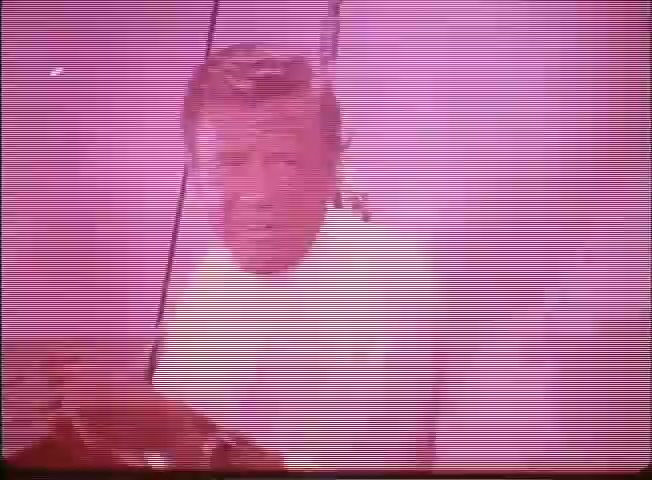 The still appears to feature a character in a white outfit, possibly an explorer or scientist, framed within a futuristic or underwater setting. The color palette leans towards a pink hue, contributing to a retro aesthetic typical of 16mm film. This frame likely emphasizes an adventurous or dramatic moment, aligning with the tone of a trailer for 'Around the World Under the Sea.' The character's expression suggests urgency or determination, fitting for a project centered on underwater exploration.