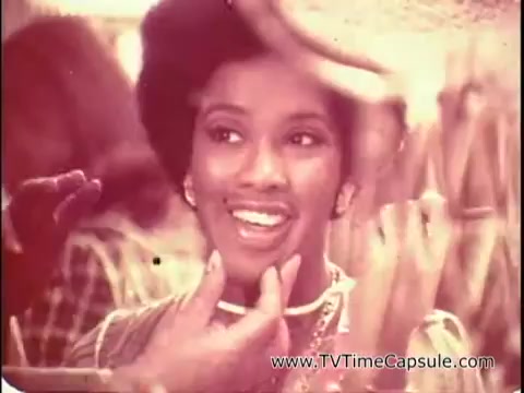 The 16mm still from 'Lasting Beauty makeup finish - 1969' features a smiling woman with a natural, glowing makeup look. She has an elegant hairstyle and is being attended to by someone off-camera, likely applying the Lasting Beauty makeup. The atmosphere seems warm and inviting, with hints of vibrant colors in the background. The emphasis is on the application process and the enhancement of her beauty, reflecting the techniques of the era in makeup artistry.