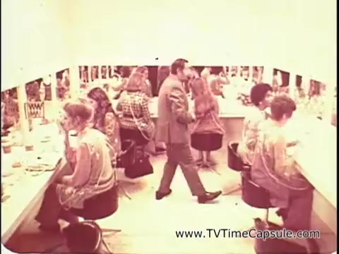 The still from 'Lasting Beauty makeup finish - 1969' showcases a vintage makeup salon scene. David Lawrence, the makeup artist, is seen interacting with clients, providing instructions as he demonstrates the makeup application process. Feminine figures, seated at makeup stations, are engaged in their beauty routines, surrounded by mirrors. The atmosphere is vibrant yet intimate, capturing a moment in time that reflects the beauty trends and cultural aesthetics of the late 1960s.