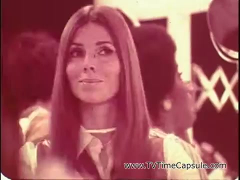 The still features a woman with long, straight hair and a gentle smile, set against a backdrop of people in a lively setting, likely a makeup demonstration or showcase. She appears to be the focus of a makeup application, highlighting the use of Lasting Beauty makeup finish by famed makeup artist David Lawrence. The ambiance suggests a 1960s vibe, capturing the era's style and beauty trends. The framing emphasizes her expression, embodying confidence and allure, which resonates with the promotion of the makeup's staying power.