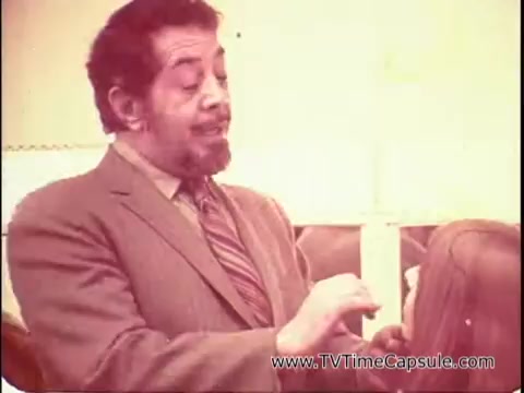 The still shows David Lawrence, a well-known makeup artist, in a professional setting, likely during a makeup demonstration. He is wearing a suit and appears engaged as he addresses an audience, instructing them to close their eyes. The environment has a vintage feel, and there seems to be a model or client receiving makeup application, highlighting the product 'Lasting Beauty makeup finish.' The overall tone suggests a focus on enhancing beauty with a professional touch.