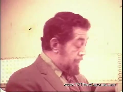 The still from 'Lasting Beauty makeup finish - 1969' shows David Lawrence, a makeup artist, focused on his work. He appears to be demonstrating a makeup application technique. The setting has a vintage aesthetic, likely featuring a makeup station with soft lighting. His expression and gestures suggest he is guiding viewers, possibly preparing them for a demonstration of the Lasting Beauty makeup. The scene reflects the era's beauty standards and techniques.