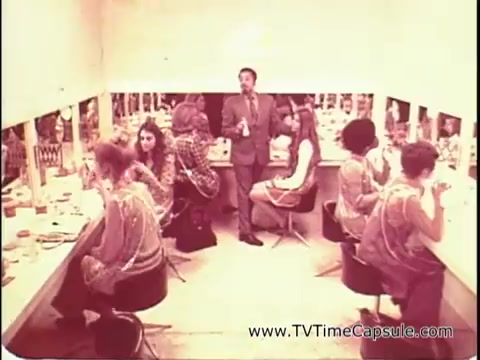 The still from 'Lasting Beauty makeup finish - 1969' features a makeup artist, David Lawrence, surrounded by several women at vanity stations. The setting appears to be a makeup studio with multiple mirrors reflecting their images. Lawrence is in a suit, demonstrating his techniques while the women, dressed in the fashion of the era, focus on their makeup application. The atmosphere is lively and professional, illustrating the beauty trends and practices of the late 1960s.