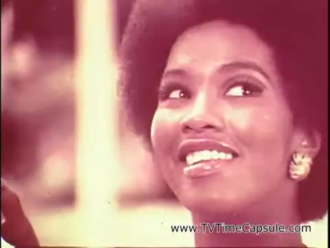 The still from 'Lasting Beauty makeup finish - 1969' features a woman with a radiant smile and an elegant afro hairstyle. She appears to be in a beauty demonstration, likely showcasing the makeup product. The warm color palette gives a vintage feel, emphasizing the era's beauty aesthetics. There seems to be a focus on her eyes and the glow of her skin, highlighting the appeal of the Lasting Beauty makeup finish as a solution for long-lasting beauty.