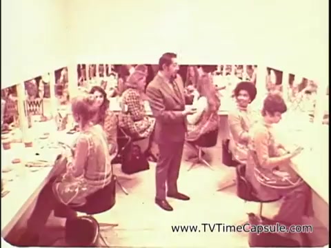 The still from 'Lasting Beauty makeup finish - 1969' features a makeup salon scene. In a bright, mirror-lined room, several women are engaging in makeup application, seated at workstations. They appear to be enjoying the process, with some looking at their reflections. David Lawrence, identified as a renowned makeup artist, stands in the center, attentively demonstrating techniques. The overall atmosphere conveys a sense of elegance and expertise in beauty enhancement during that era.