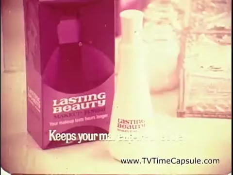The still features a promotional scene for 'Lasting Beauty' makeup finish from 1969. The image showcases a bottle of the product, which is white with the brand name prominently displayed. Next to it sits a purple box with the same branding. The tagline 'Keeps your makeup looking fresh longer' hints at its purpose. The aesthetic is retro, typical of the era, with a warm color tone. In the background, there are hints of a glamorous makeup setup, suggesting a focus on enhancing beauty.