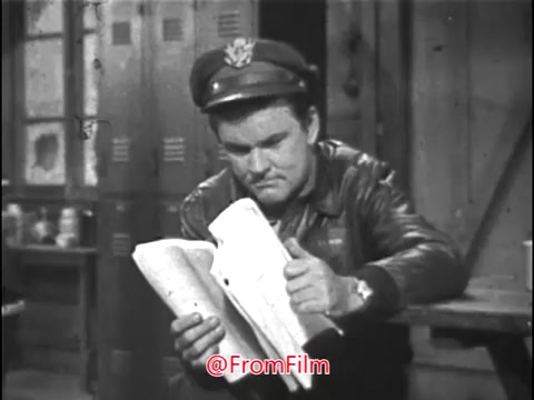 In this 16mm still from a Public Service Announcement (PSA) featuring Bob Crane, he is dressed in a military flight jacket and hat, indicating his role as a pilot. The scene appears to be set in a military environment, likely a barracks, as suggested by the lockers in the background. Bob Crane is engaged in reading a document, looking focused and serious, which aligns with the message he is about to deliver about the important work of experienced pilots. The off-screen voice prompting 'Action!' indicates the filming in progress. The overall tone suggests a blend of entertainment with a significant message about service in the Air National Guard.