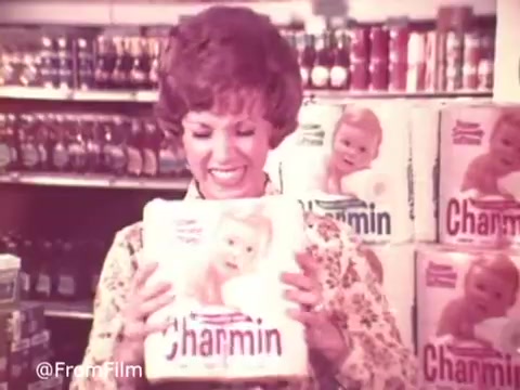 The still from the 1970s Charmin commercial features a cheerful woman holding a package of Charmin toilet paper with a baby on the cover. She has a big smile, conveying happiness and satisfaction with the product. In the background, shelves are stocked with various grocery items, adding to the supermarket ambiance. The commercial captures the lighthearted and humorous spirit of the era, famously associated with Mr. Whipple's warning: 'Don't squeeze the Charmin!'